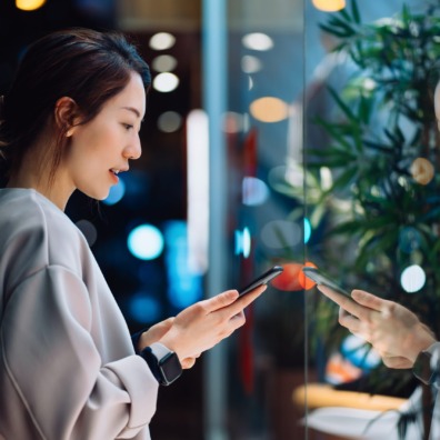 Smiling young Asian woman using smartphone while shopping in the city, standing outside a boutique shop window in the evening